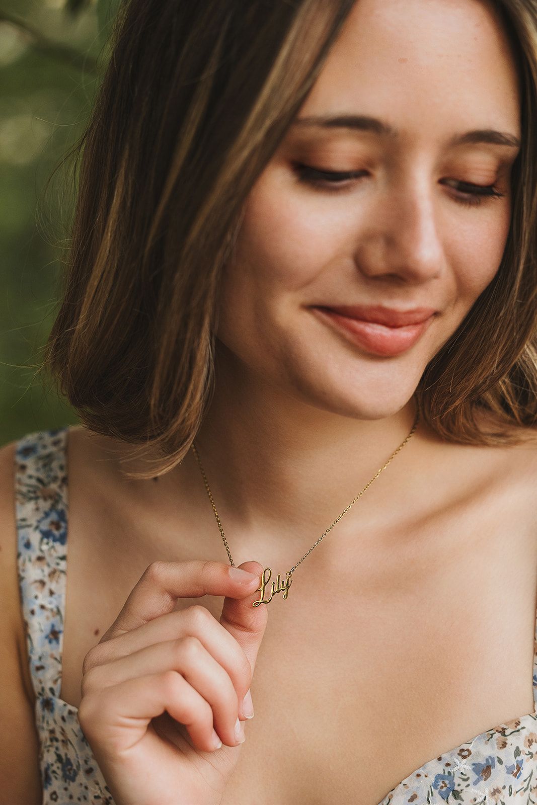 Close-up portrait of someone looking down with a gentle smile while holding a delicate gold necklace pendant.