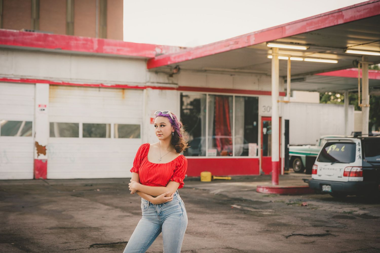 Person in a red crop top and high-waisted jeans poses outside a retro-style gas station in warm lighting.