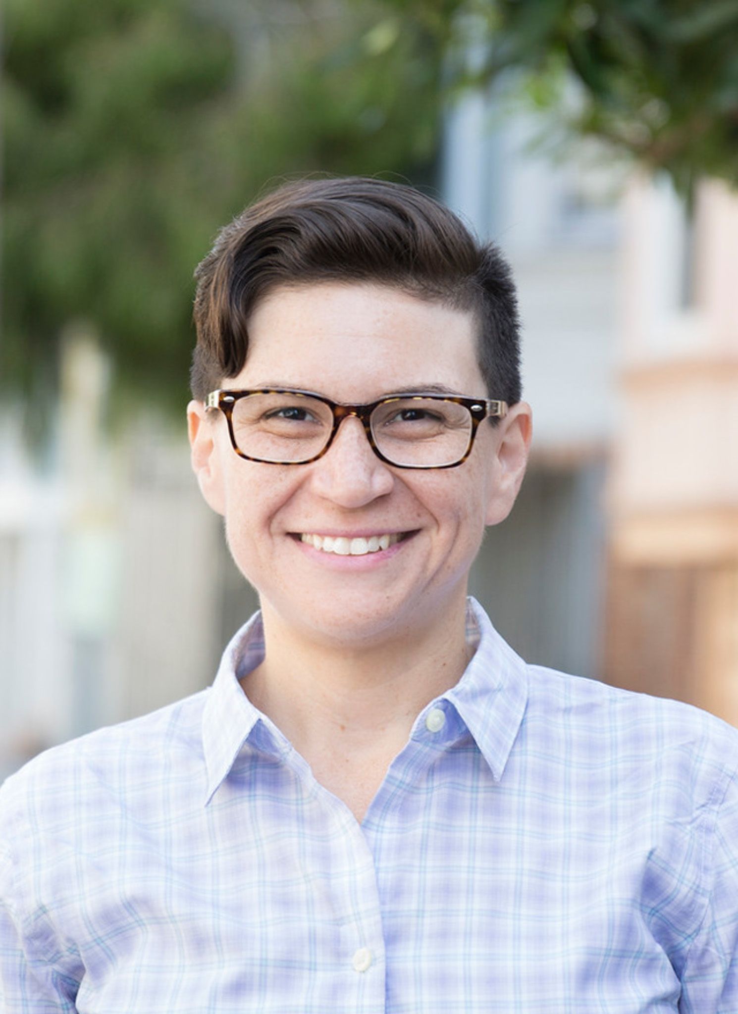 A smiling person with glasses and well-styled hair, wearing a light blue checkered shirt, standing in a colorful alley.
