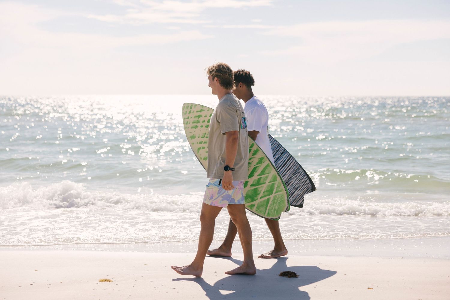 Two people in casual beachwear walking along a sunny shoreline with surfboards.
