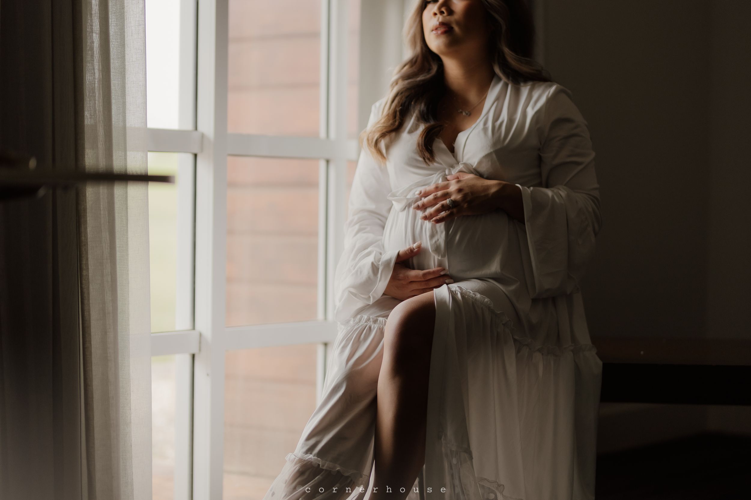 A person in a light-colored dress sits on a bed in a room with natural light streaming through windows.