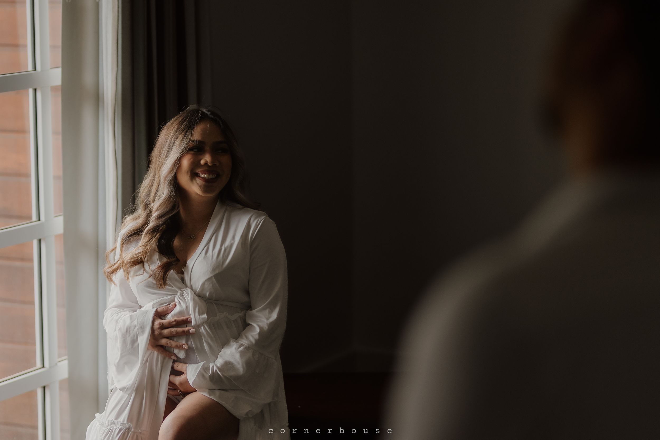 Person in white robe sits by window with soft natural light creating moody boudoir atmosphere.