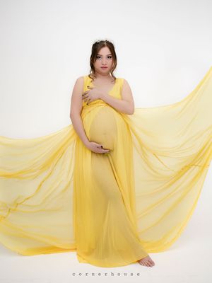 Figure in flowing yellow chiffon maternity gown stands against white backdrop as fabric billows dramatically.