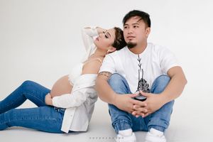 A couple wearing white tops and blue jeans sit and recline on light backdrop in casual, intimate poses.