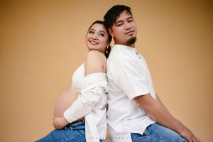 A couple in white outfits poses back to back against a warm brown background in a casual studio portrait.