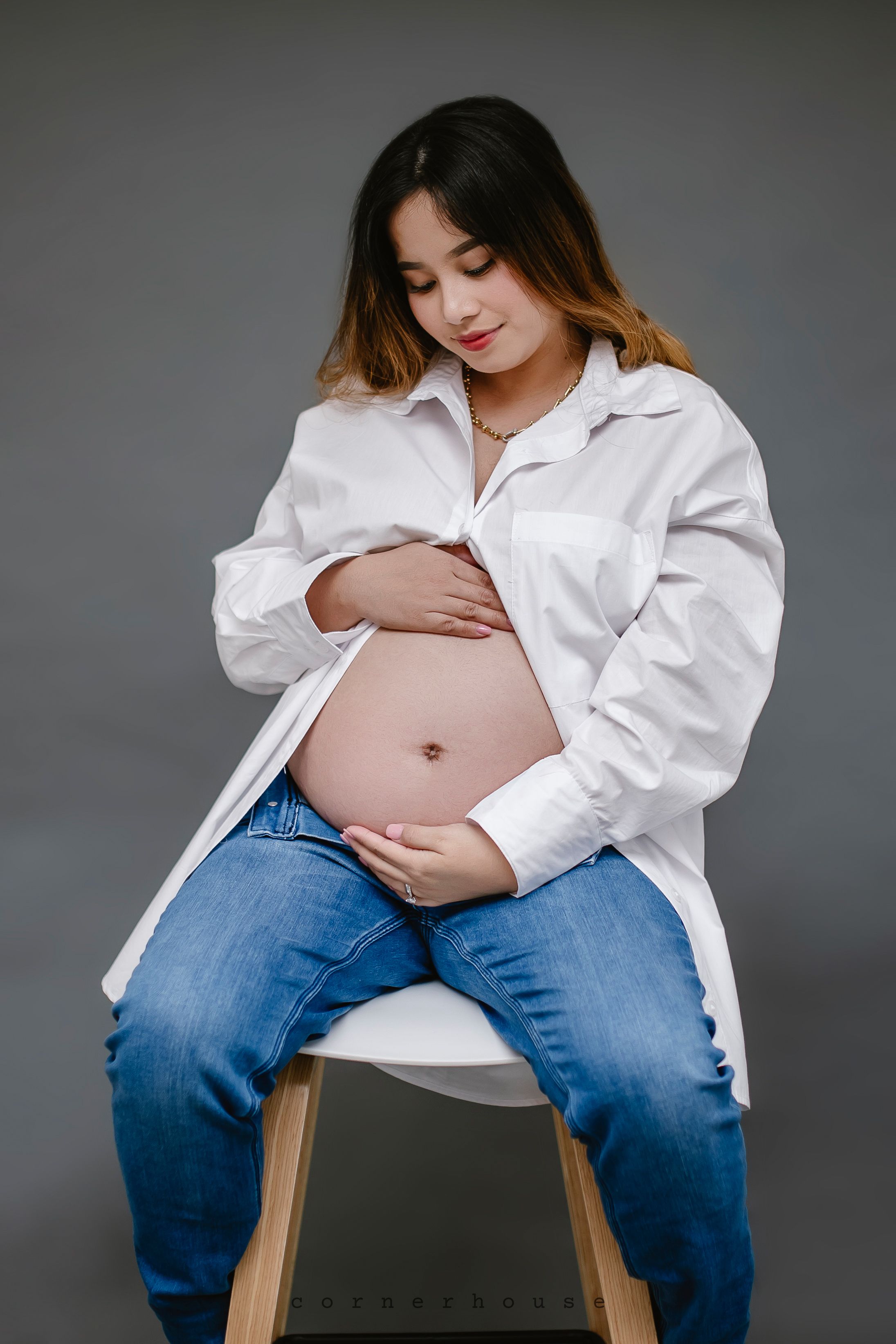 Pregnant person in white shirt and jeans sitting on stool, touching belly with downward gaze against gray background.