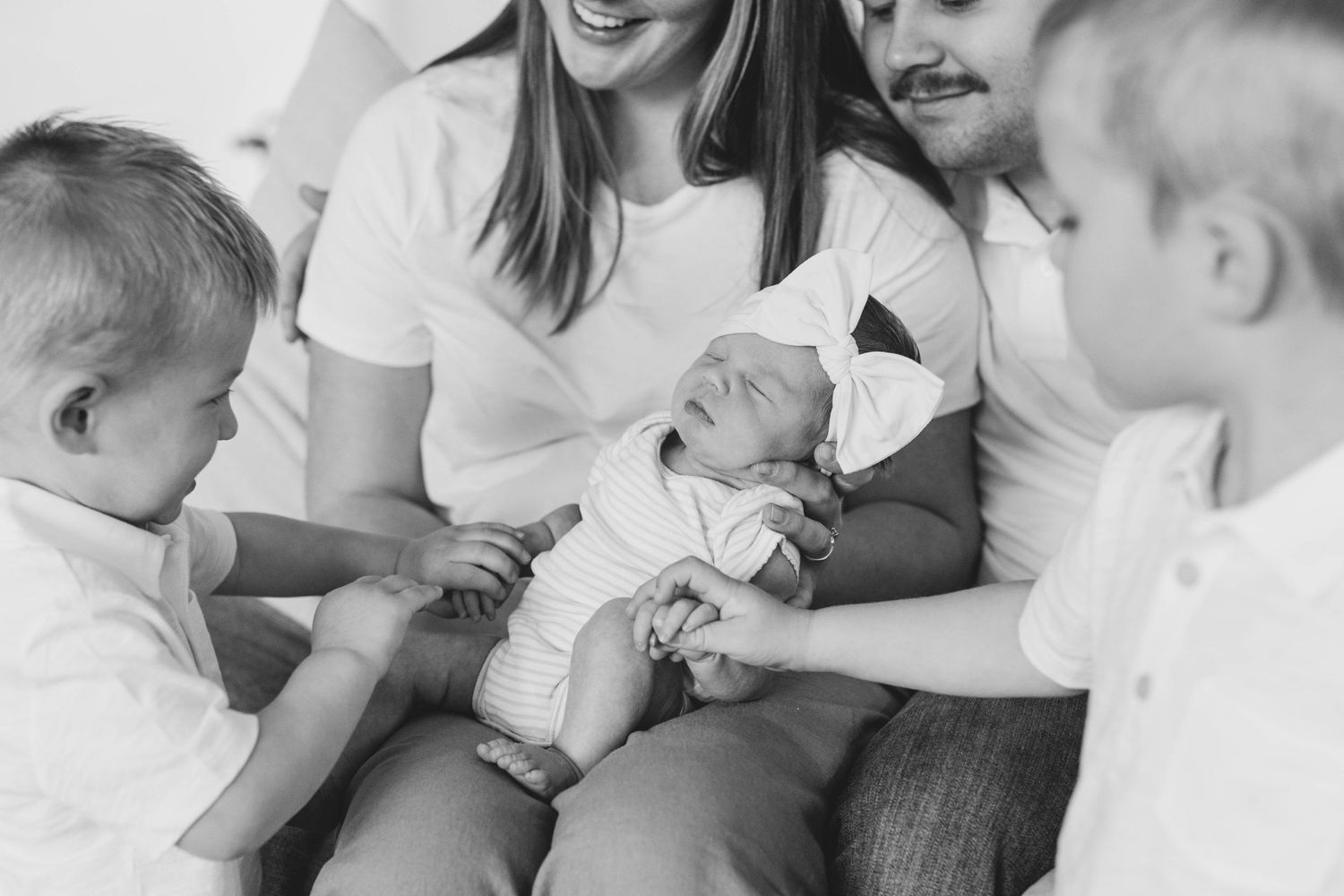 A tender black and white family moment showing a newborn baby being gently touched by two young siblings in white.