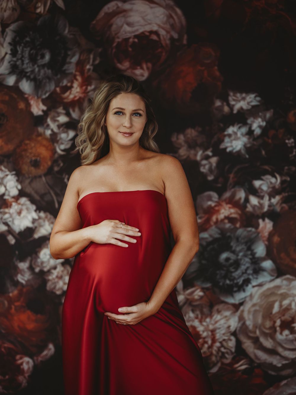 A maternity photoshoot featuring a red silk dress against a dark floral backdrop creates an elegant and dramatic atmosphere - Bellevue WA