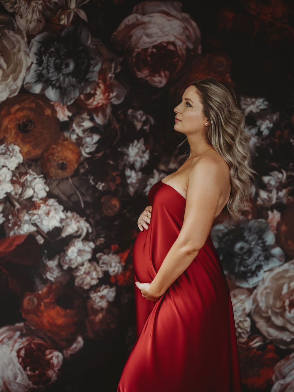 A woman in a flowing red maternity dress poses against a dark floral backdrop in an artistic photoshoot - maternity photography Bellevue WA