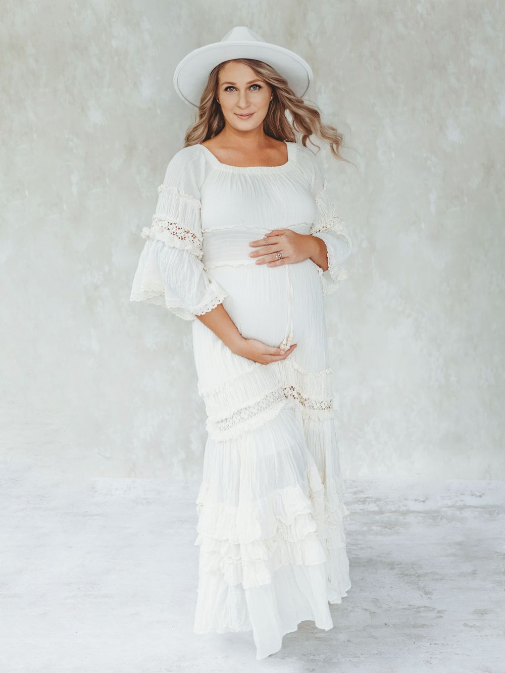 Woman in flowing white maternity dress poses against light gray backdrop in elegant pregnancy photoshoot maternity photography Bellevue WA