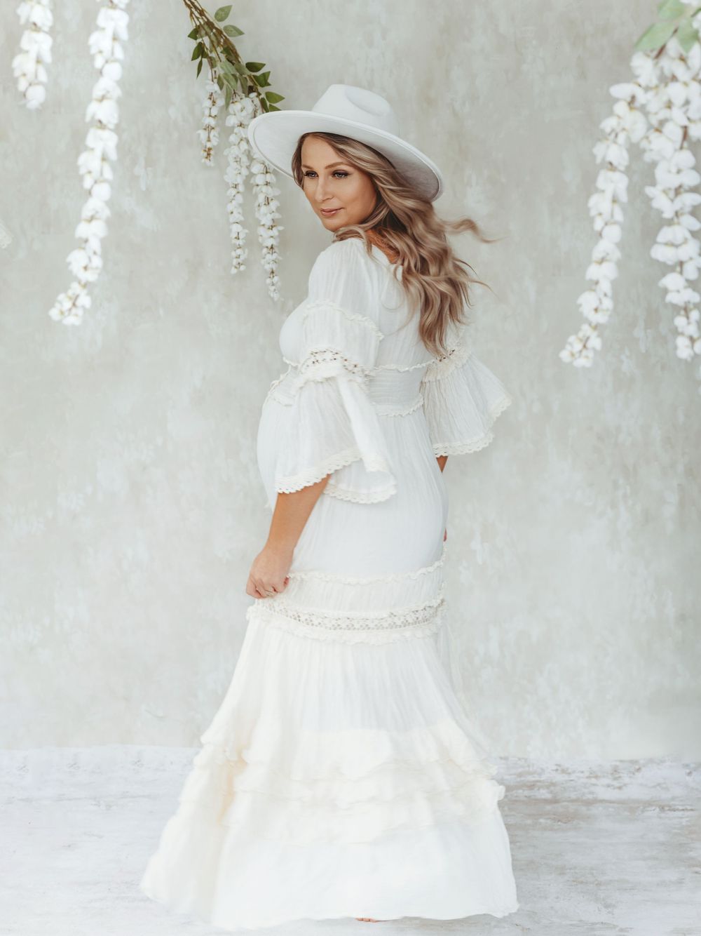 Ethereal bridal portrait featuring a white flowy dress with bell sleeves and wide brim hat against hanging floral backdrop - maternity photo