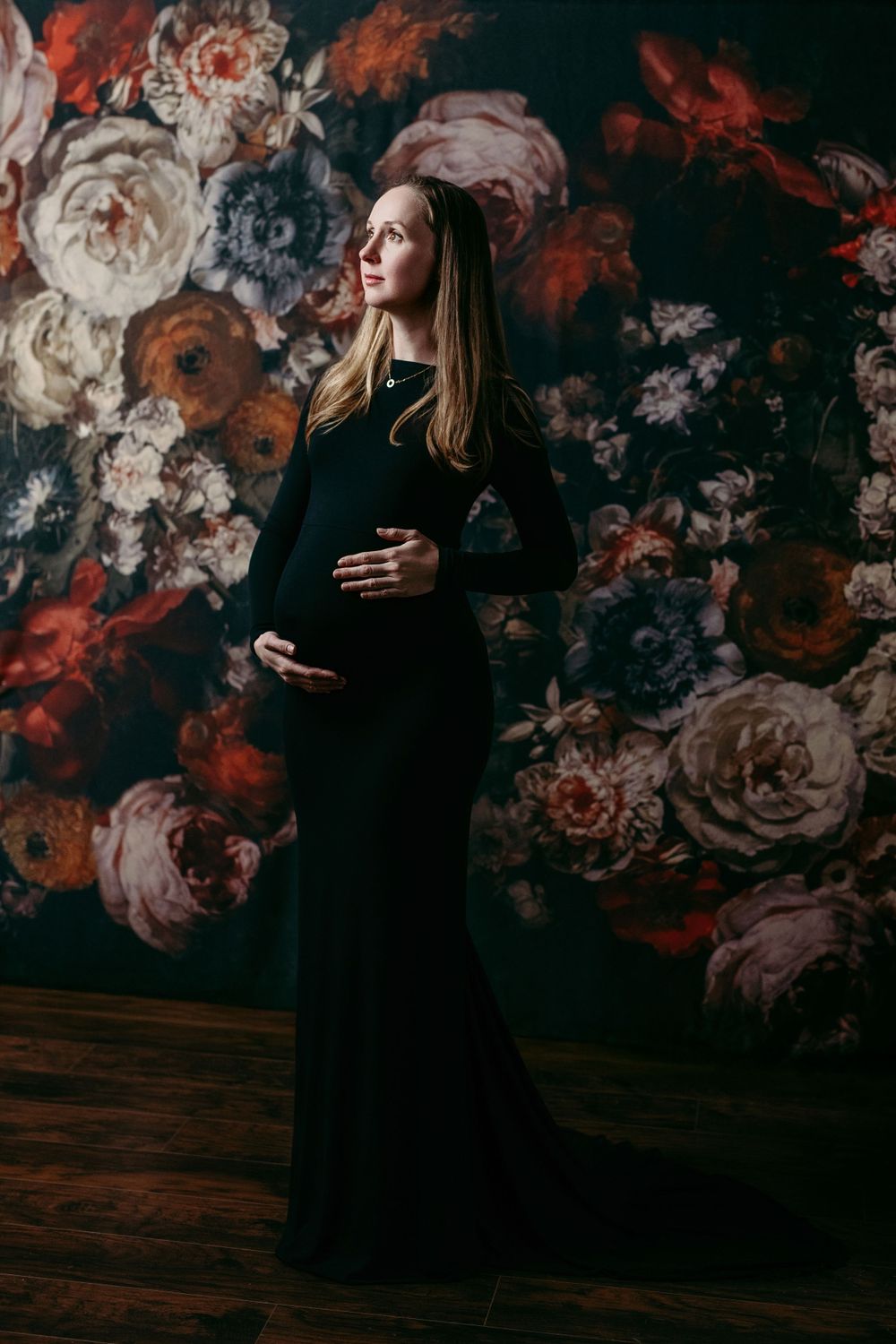 Maternity portrait against dark floral wallpaper featuring elegant black dress and dramatic lighting. Bellevue studio, WA