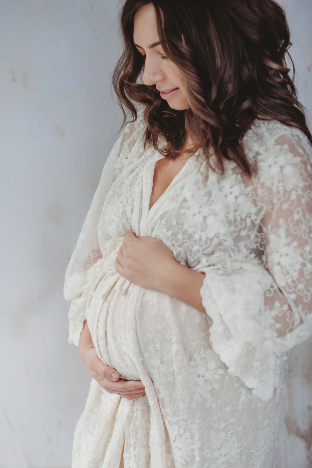 Pregnant woman in delicate white lace robe gently cradles her baby bump against light background in Bellevue studio.
