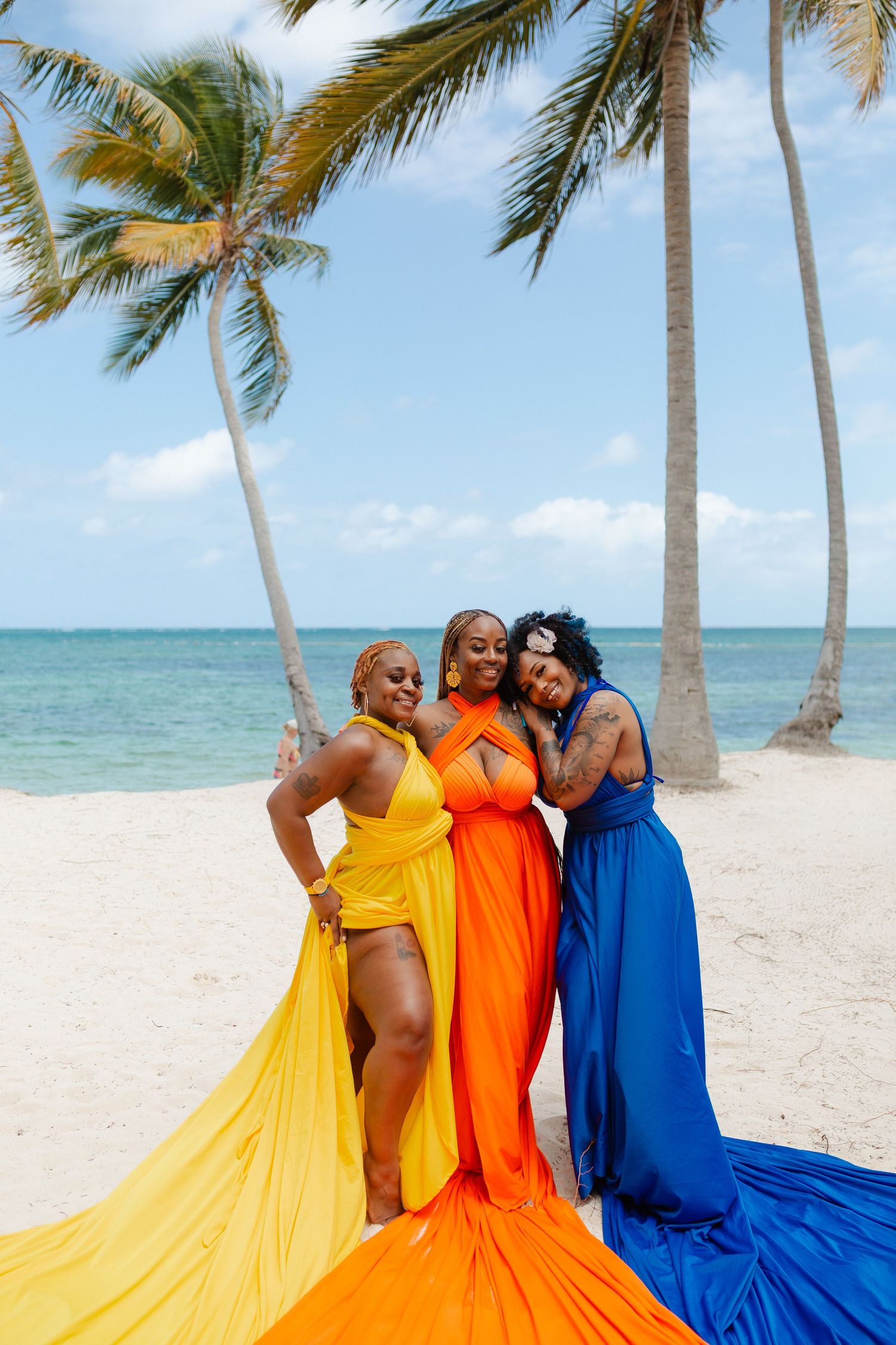 Three flowing dresses in yellow, orange and blue colors swirl on a tropical beach with palm trees and turquoise waters.