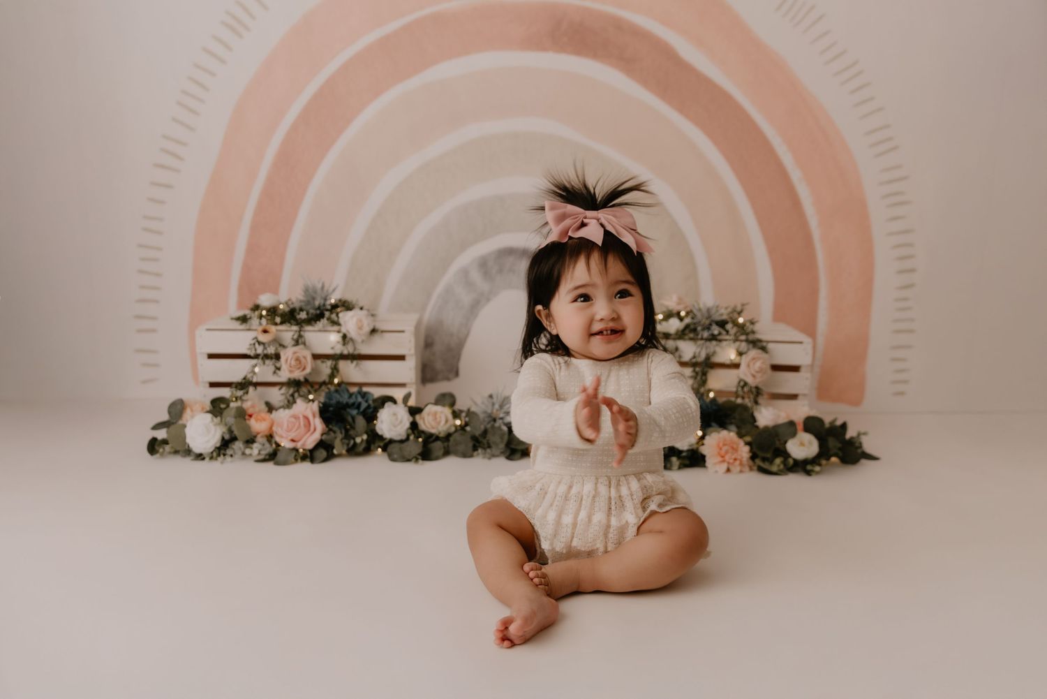 Baby in white romper sits in front of rainbow arch backdrop with flowers and books in studio setting.