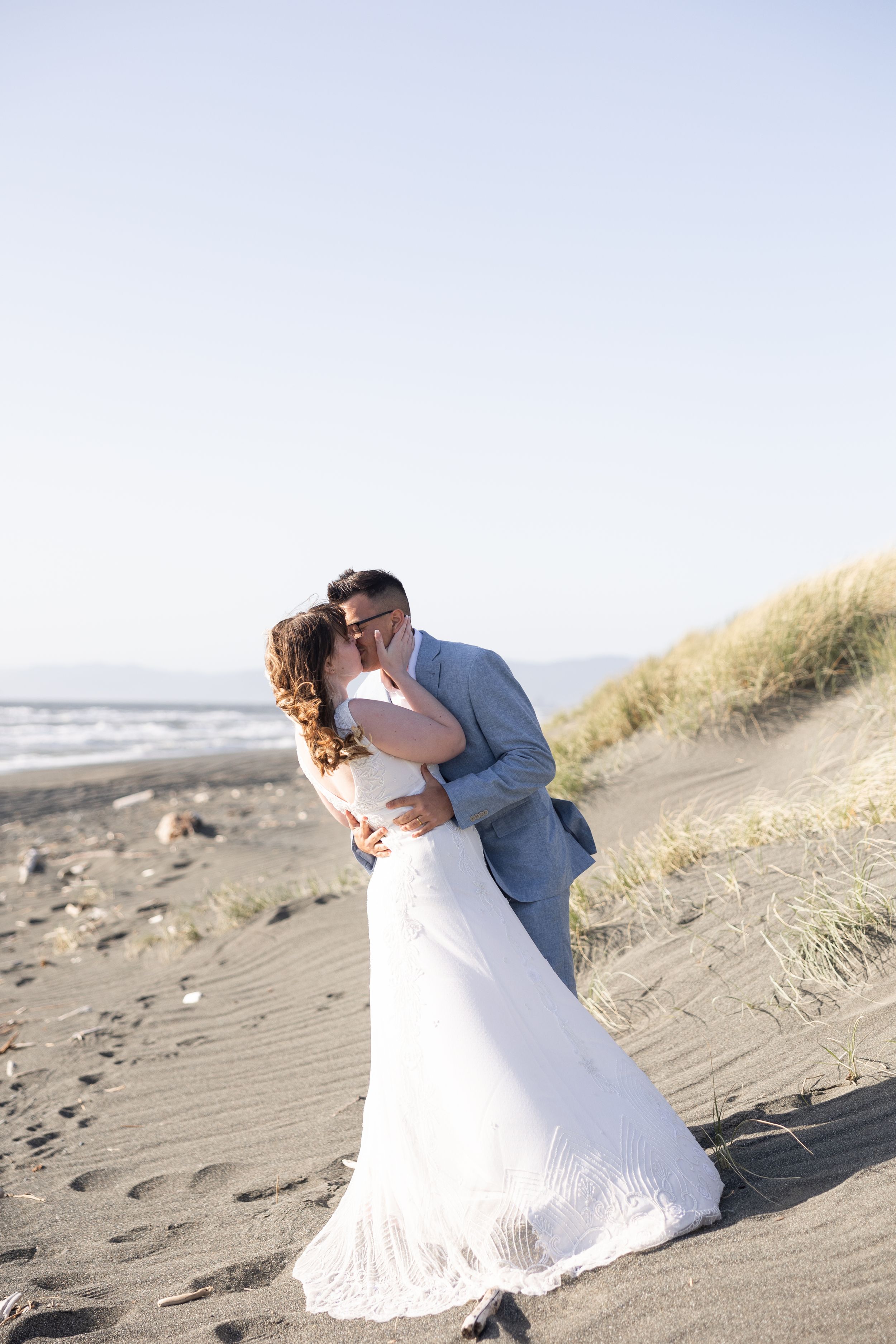 Newlyweds kissing on sandy beach at sunset in wedding attire.