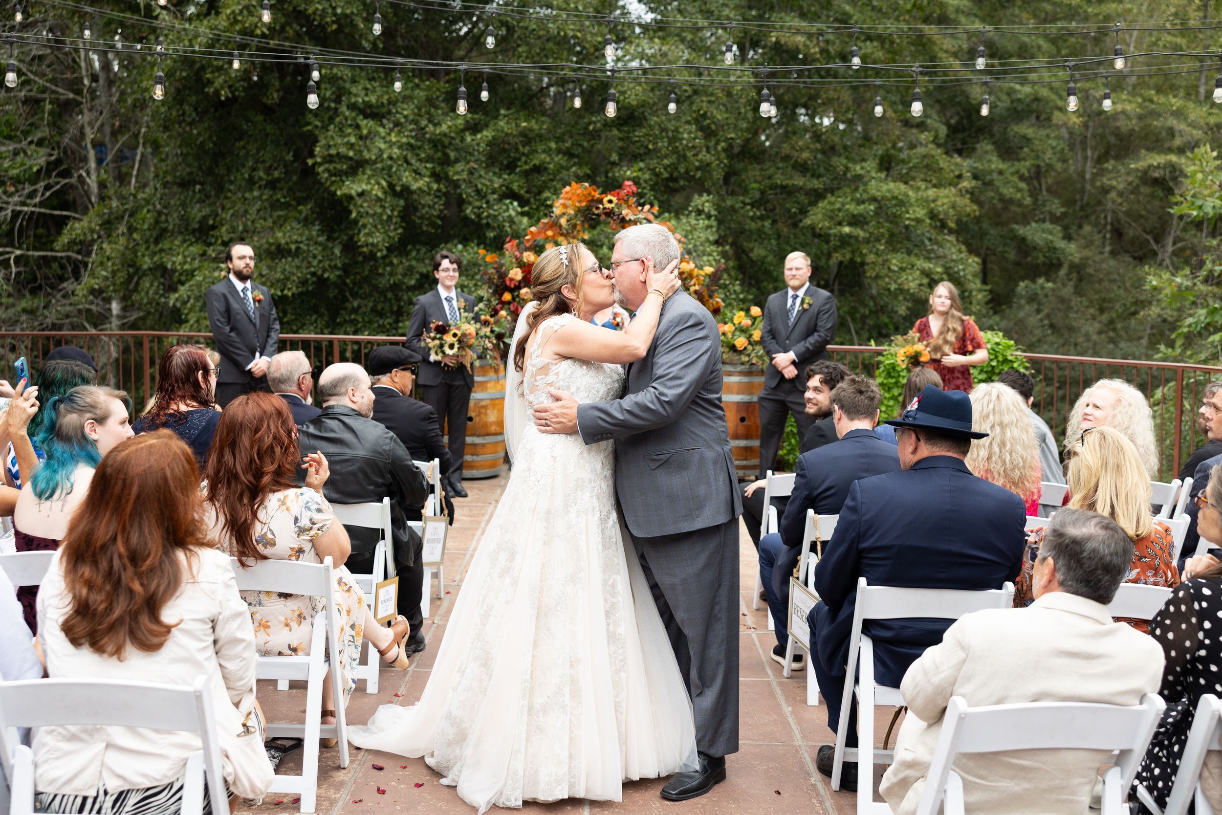 Bride and groom share first kiss at outdoor wedding ceremony with string lights overhead.