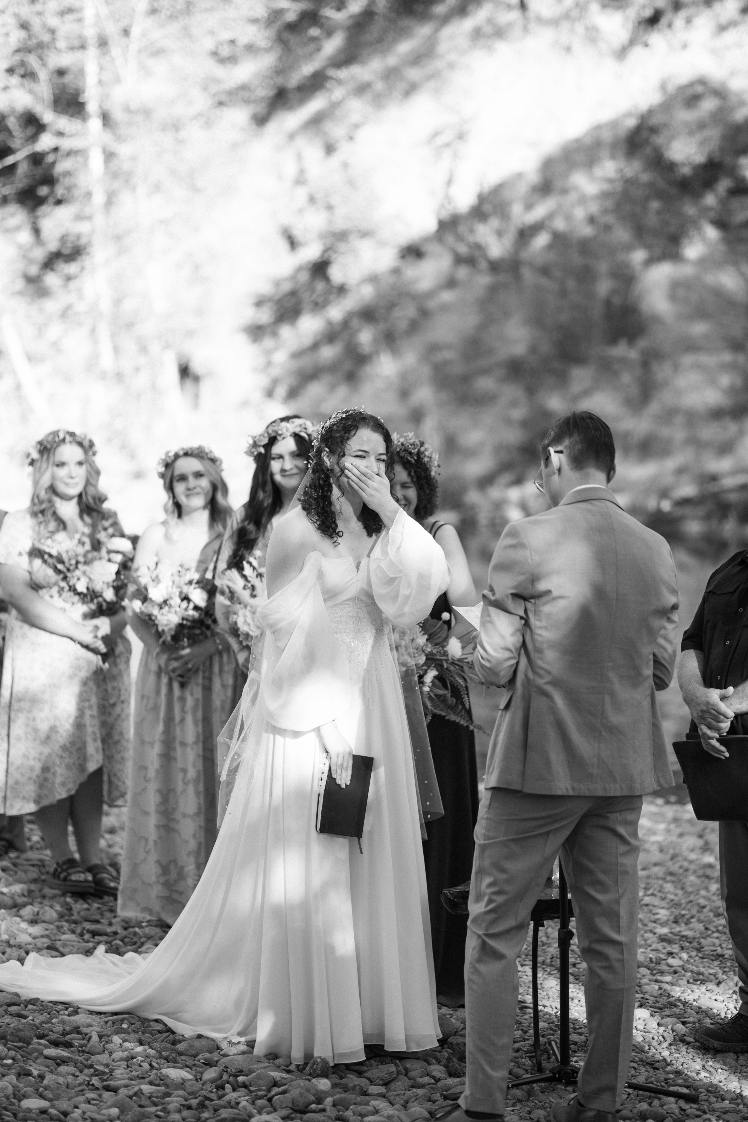 Emotional bride crying during outdoor wedding ceremony with bridesmaids in floral crowns standing behind her.
