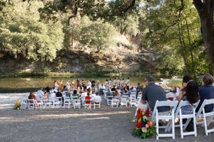 Outdoor riverside wedding ceremony with white chairs, guests seated, and colorful floral arrangements in natural setting.