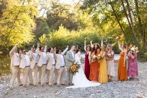Wedding party celebrating on rocky riverbank with colorful bridesmaid dresses and lush green trees in background.