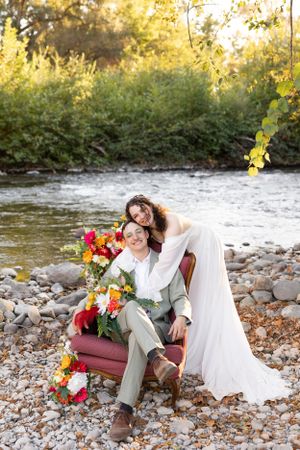 Bride and groom posing by a river on rocky shore with colorful floral arrangement and autumn foliage backdrop.