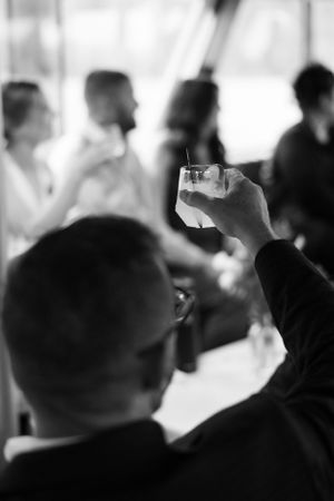 Black and white photo of a man raising a glass of whiskey at a social gathering with blurred guests in background.