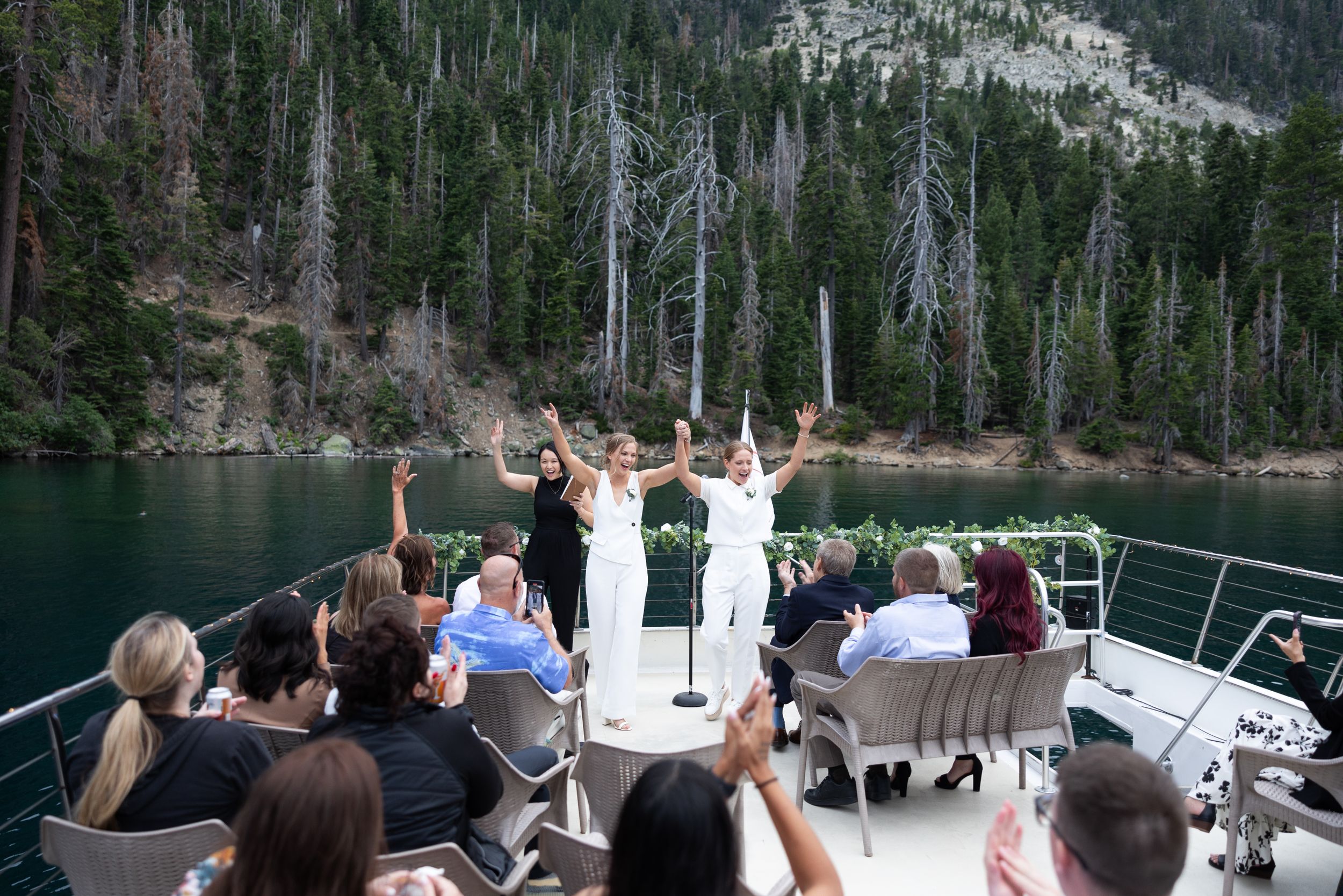Two brides in white celebrate on a boat deck with guests and a forested lake backdrop.