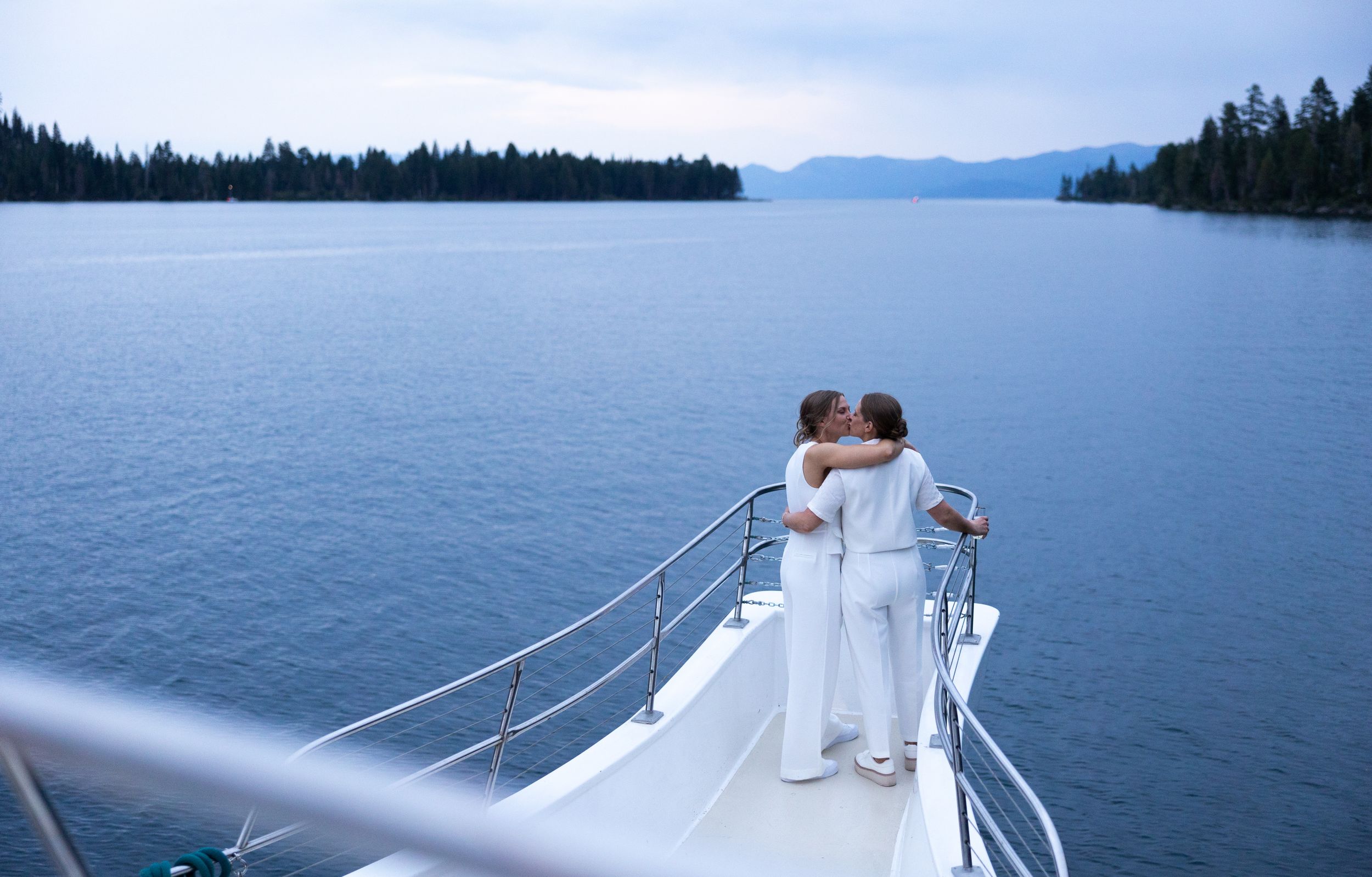 Two brides in white share a kiss on the bow of a boat on a serene blue lake.