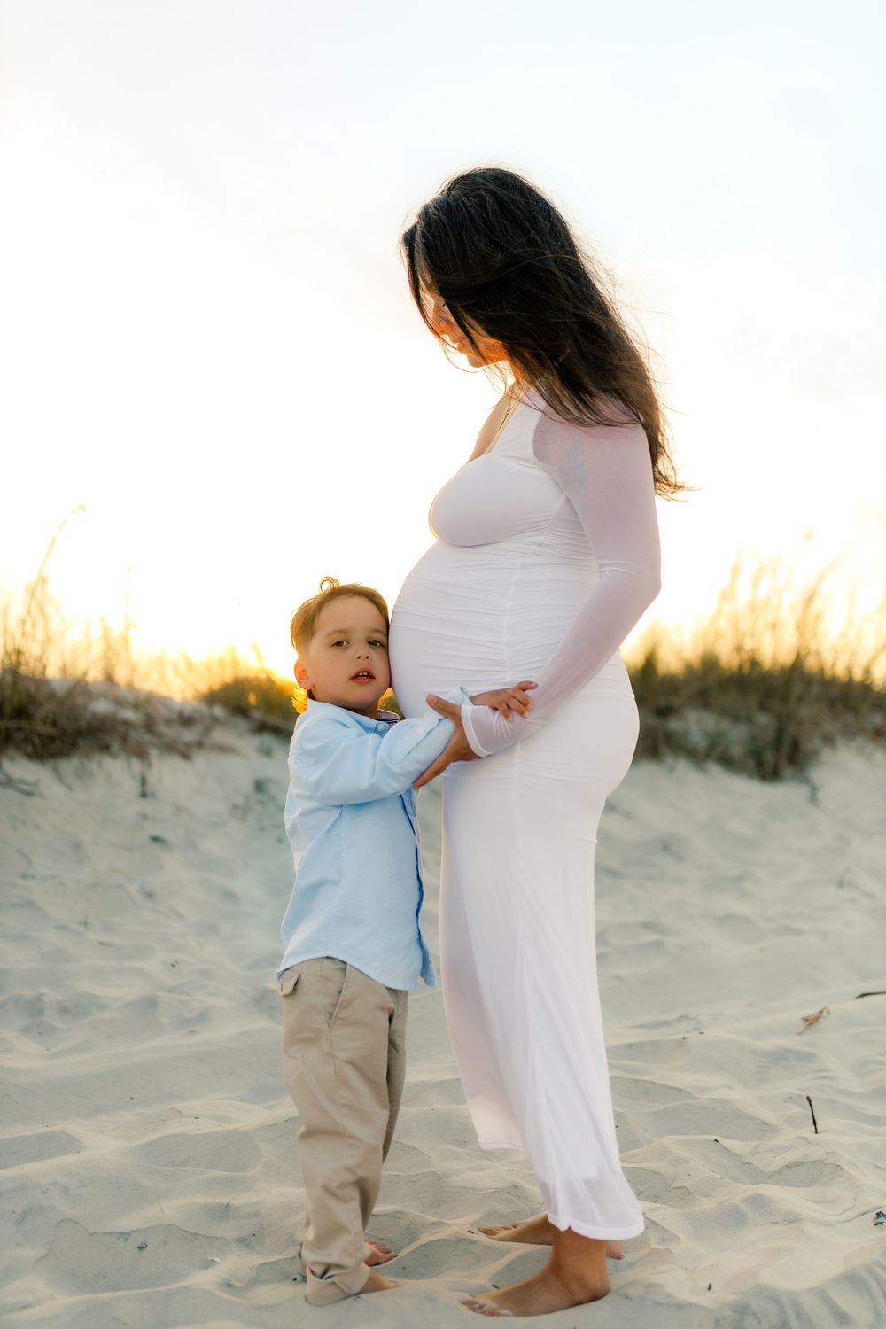 A young boy touches his pregnant mother's belly on a sandy beach at sunset, both dressed in light colors.