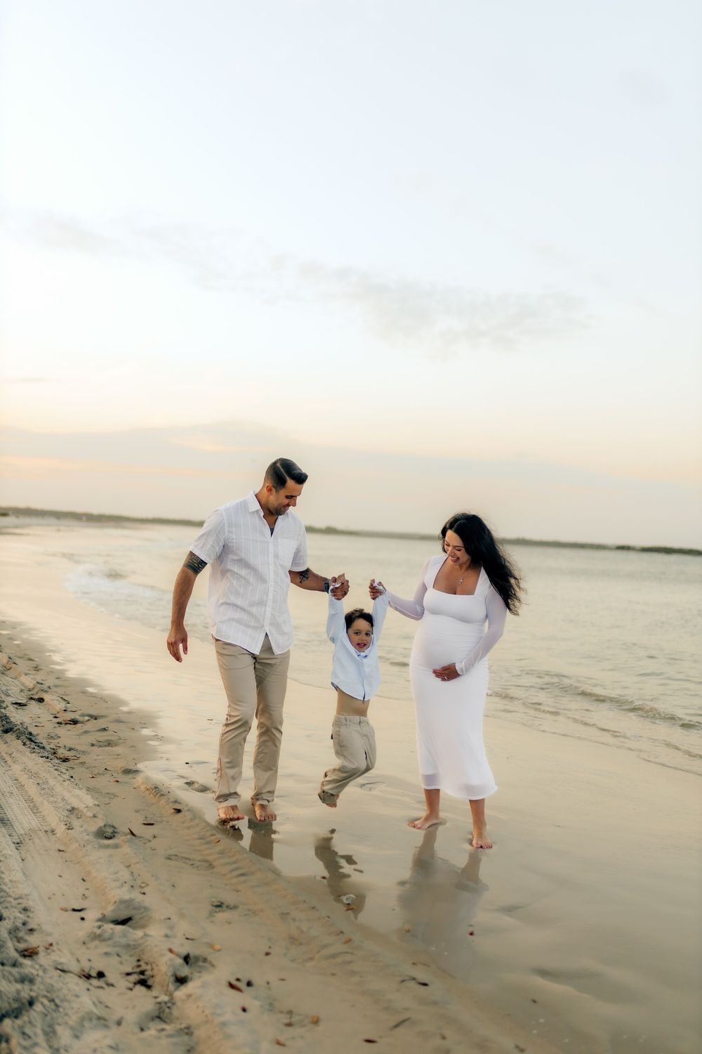 A pregnant woman in a white dress walks on the beach with her partner and young child at sunset.