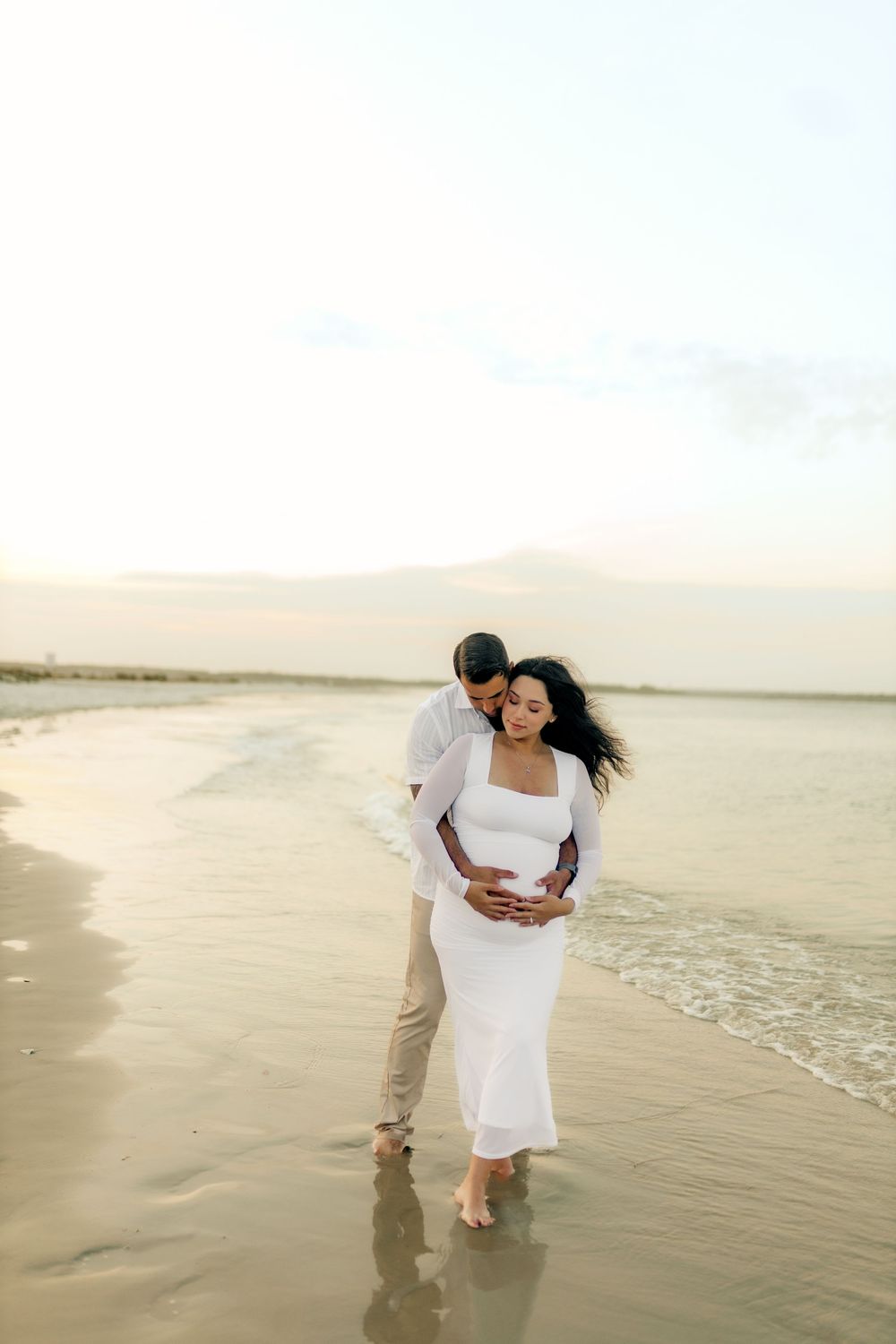 A man tenderly embraces his pregnant partner in a white dress on a serene beach at sunset.