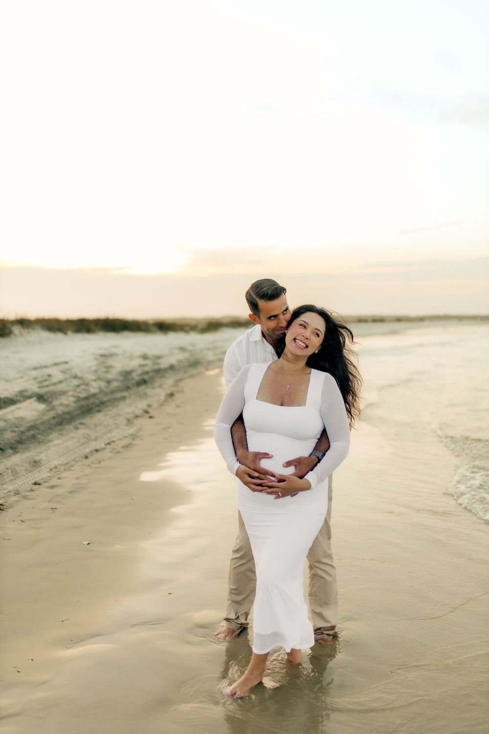 Couple embracing on beach at sunset, pregnant woman in white dress smiling, man holding her belly near ocean shore.