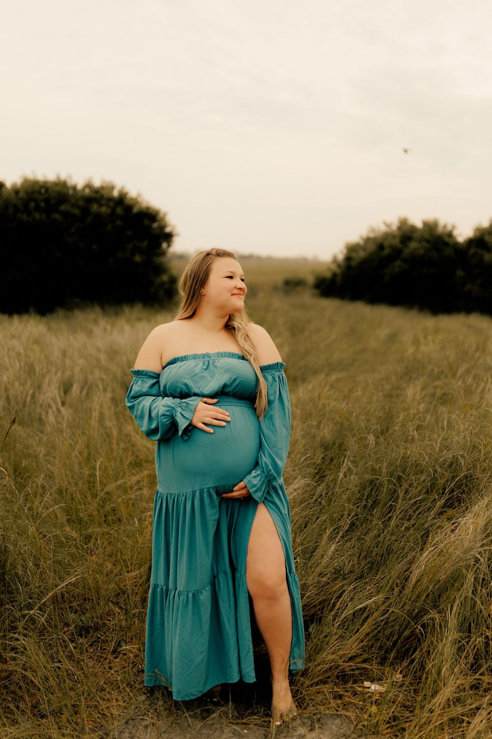 Pregnant woman in teal off-shoulder gown cradling baby bump in lush green field.