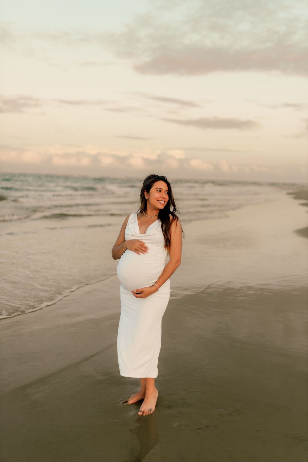 Pregnant woman in white dress cradling baby bump on beach at sunset.