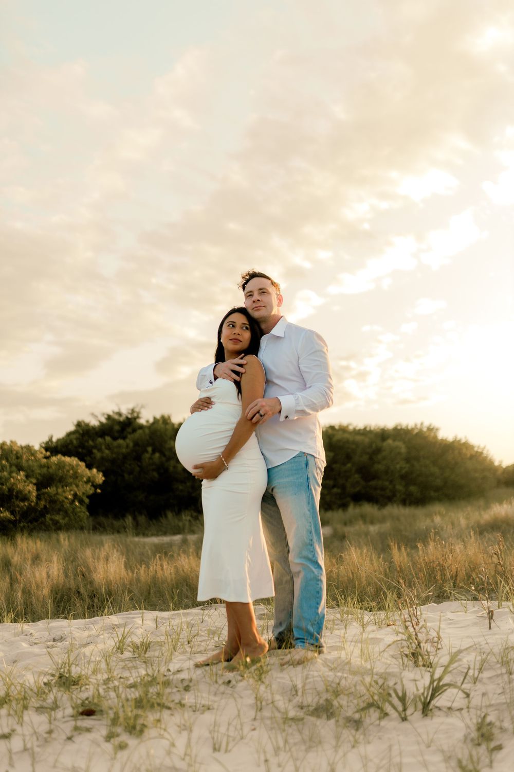 Pregnant couple embracing on sandy dunes at sunset, woman in white dress cradling bump, man in white shirt and jeans.