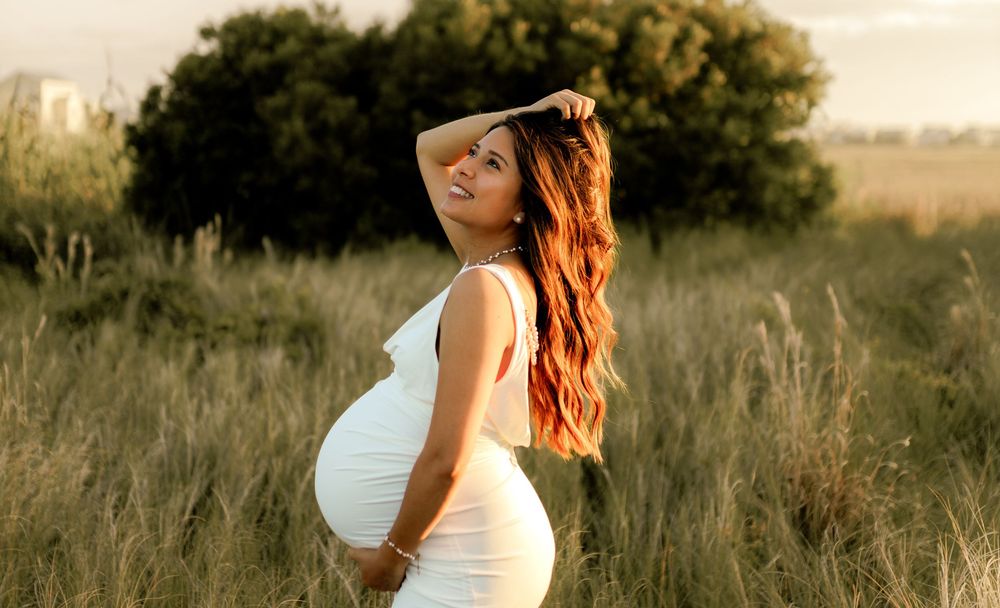 Pregnant woman in white dress posing in golden field at sunset with long red hair and glowing skin.