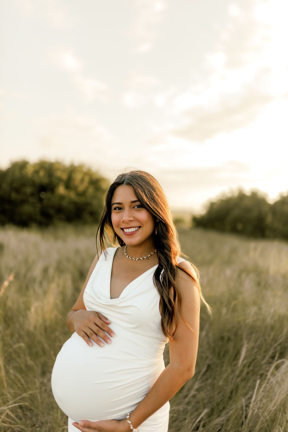 Smiling pregnant woman in white dress holding baby bump in golden field at sunset.