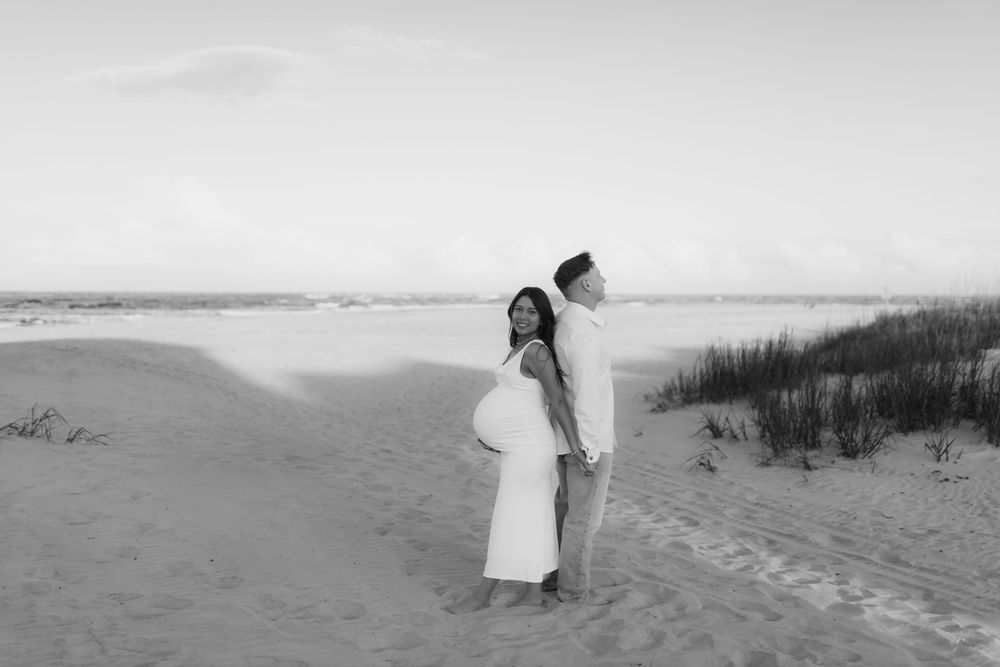 Black and white maternity photo of pregnant couple standing back-to-back on sandy beach dunes.
