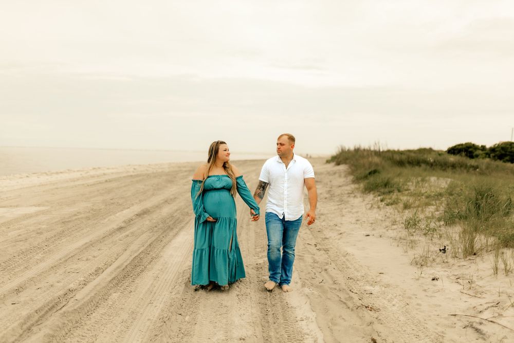 Pregnant couple holding hands walking on a sandy beach path, woman in teal maternity gown, man in white shirt.