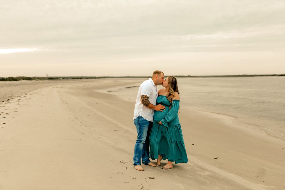 Couple embracing on sandy beach, woman in teal maternity gown, man in white shirt and jeans, cloudy sky backdrop.