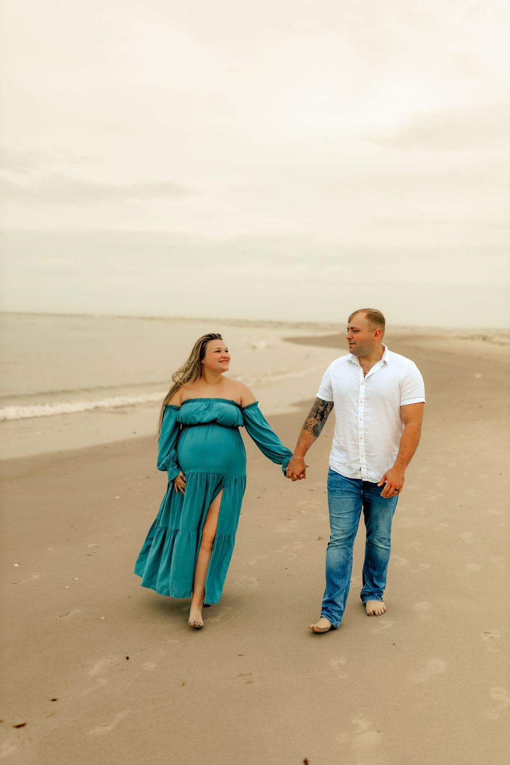 Expecting couple holding hands walking barefoot on beach, woman in teal maternity gown.