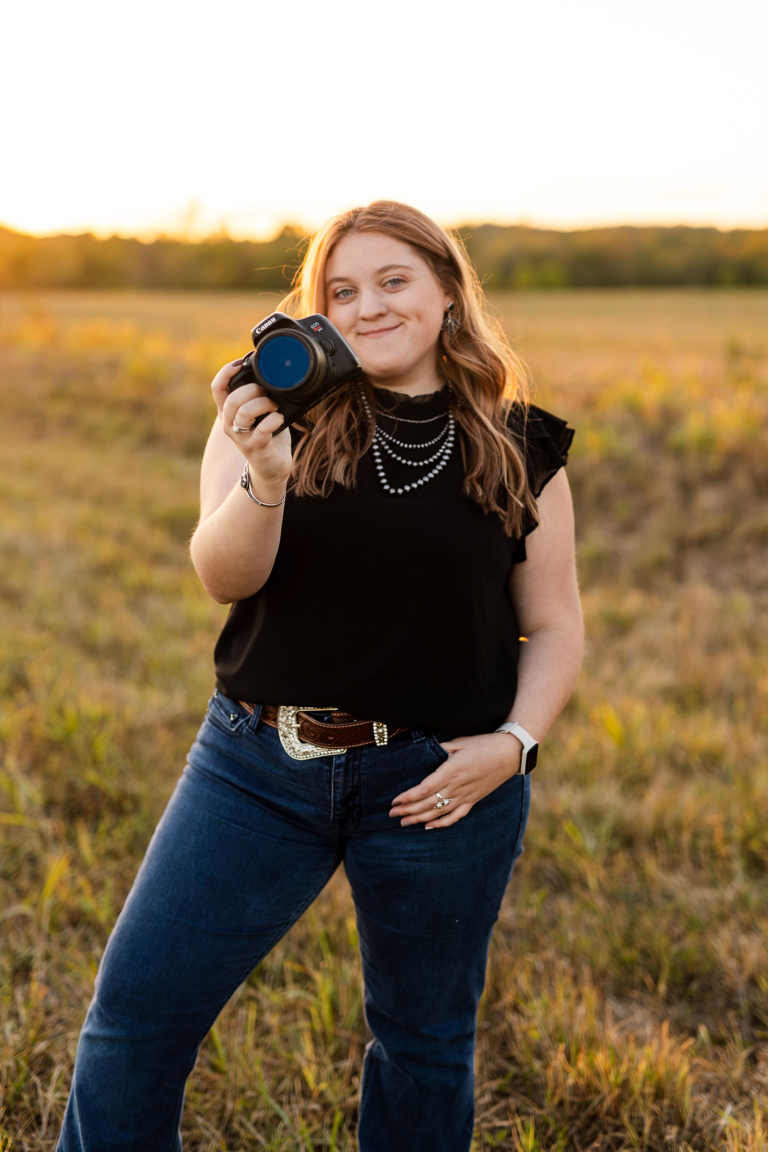 Laura- Smiling woman photographer holding a camera in a sunlit field at golden hour sunset.