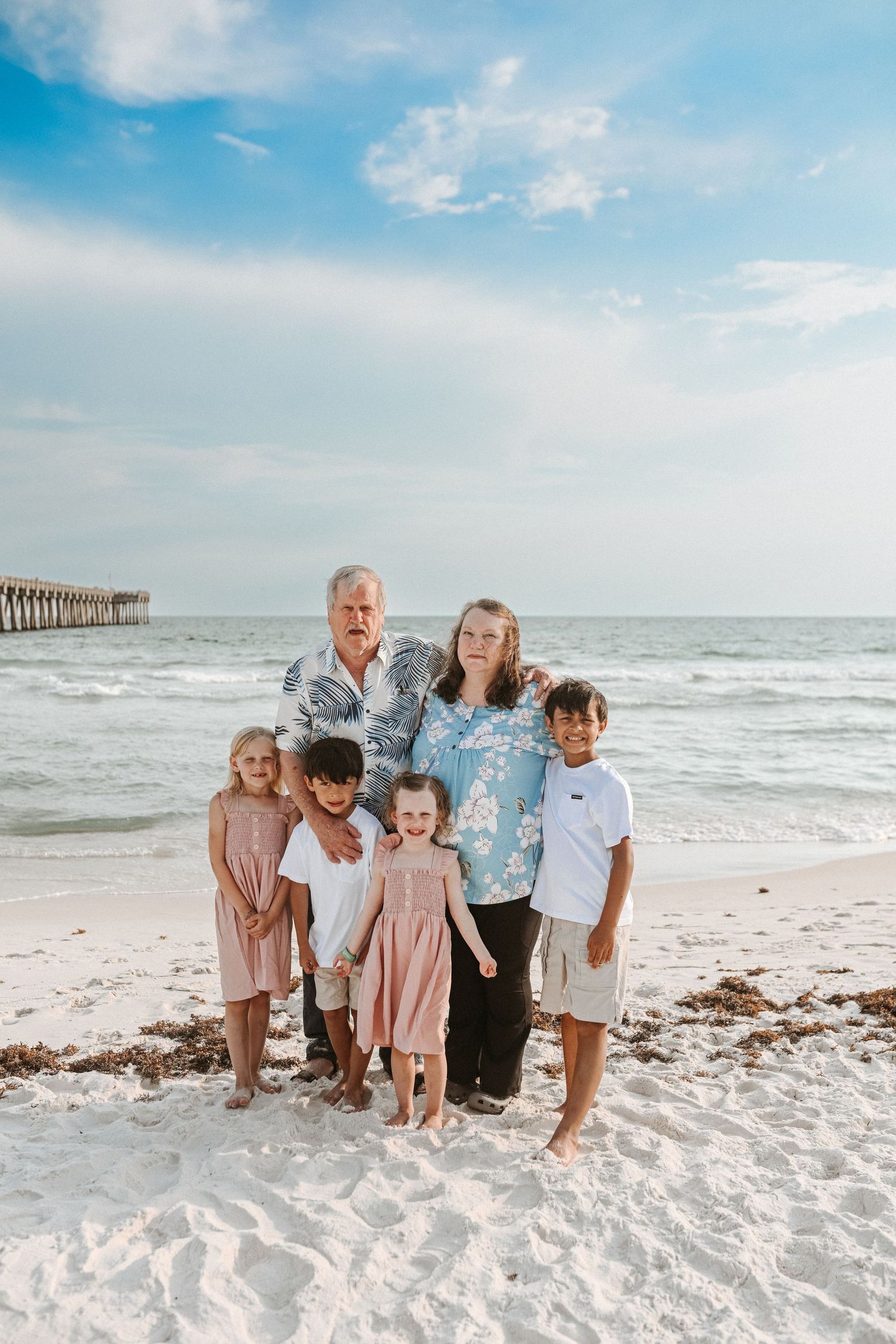 Family group poses together on white sandy beach with ocean waves and pier in background.