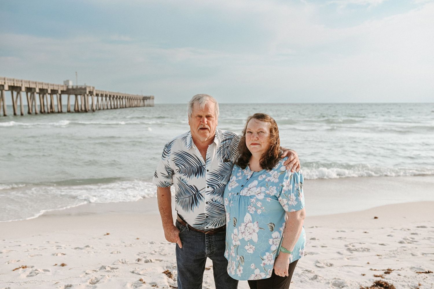 Family members embrace while standing on beach with wooden pier visible in distance.