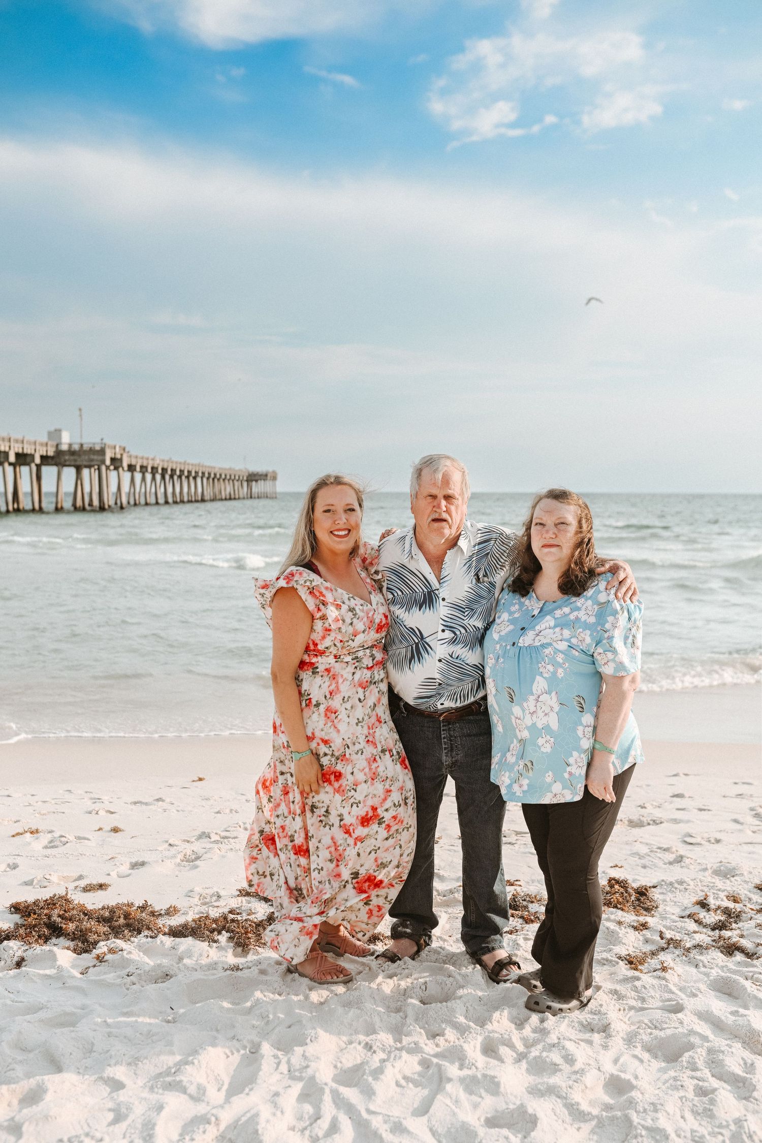 Family members pose for casual beach portrait with ocean waves and pier behind them.
