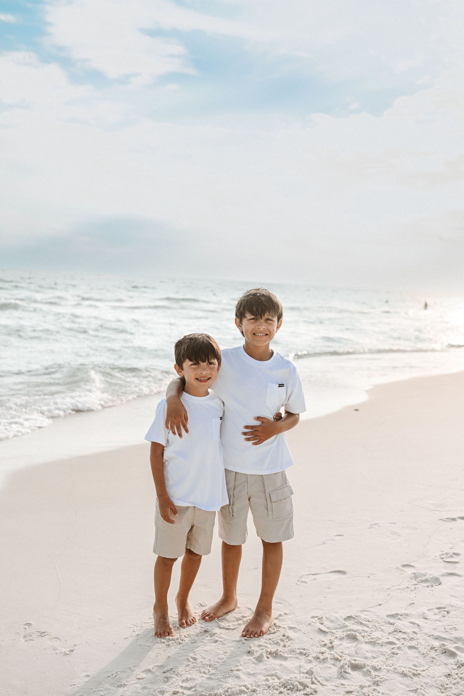 Two children in white outfits stand together on sandy beach.