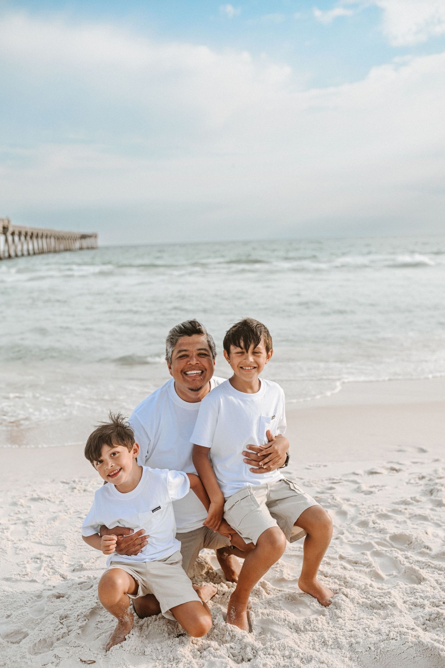 A family enjoys a playful moment together on a white sandy beach with ocean waves in the background.
