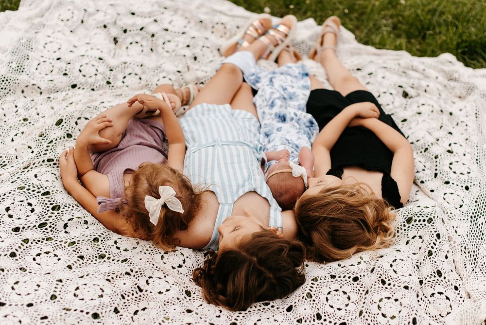 Four young sisters laying on a white crochet blanket, cuddling their newborn sister. Lindsay Miller Photography