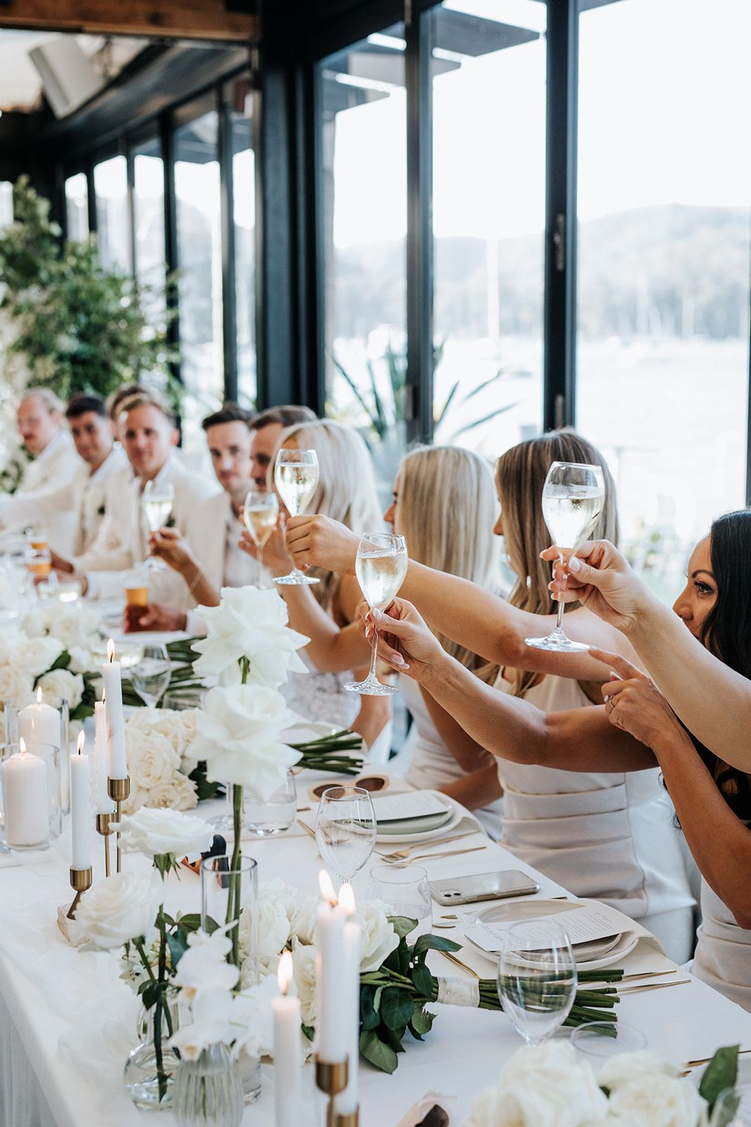 Wedding guests raise champagne glasses in a toast at elegant white floral reception table.