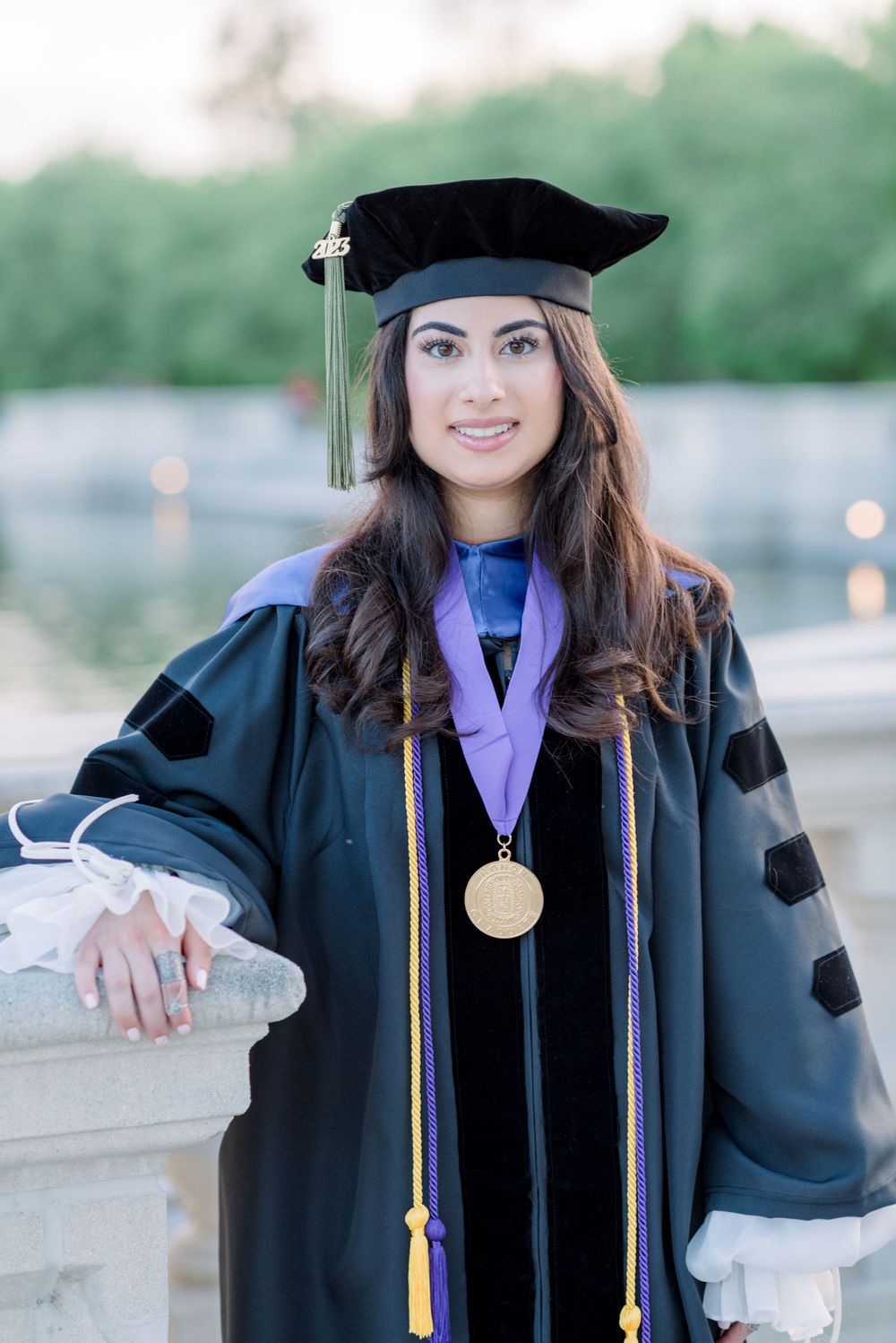 Graduate in traditional doctoral cap and gown with purple sash and gold medallion poses outdoors during commencement.